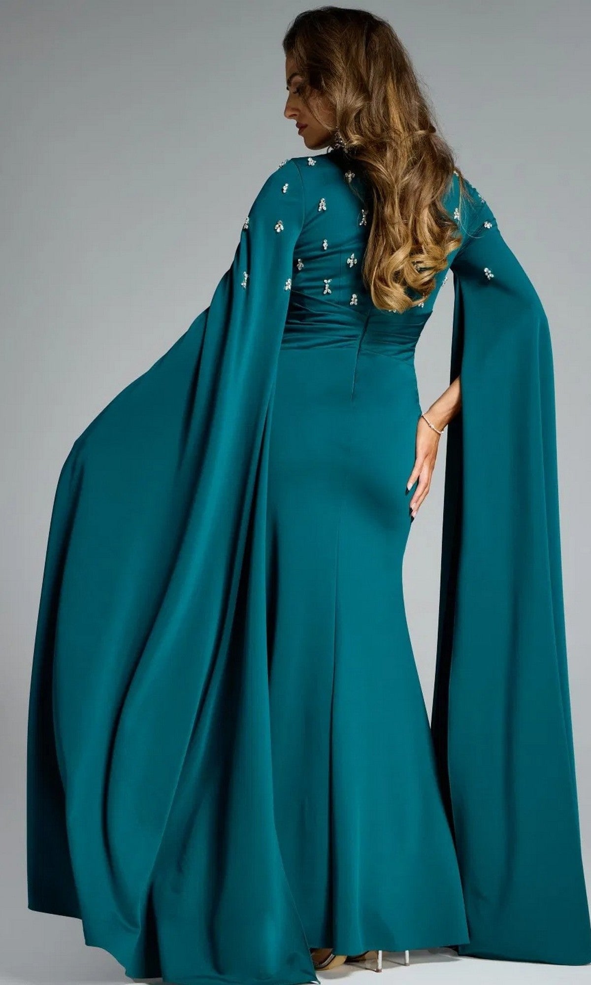 Woman showing the back of a dark green evening gown with cape-style sleeves that she is wearing, standing in front of a gray background